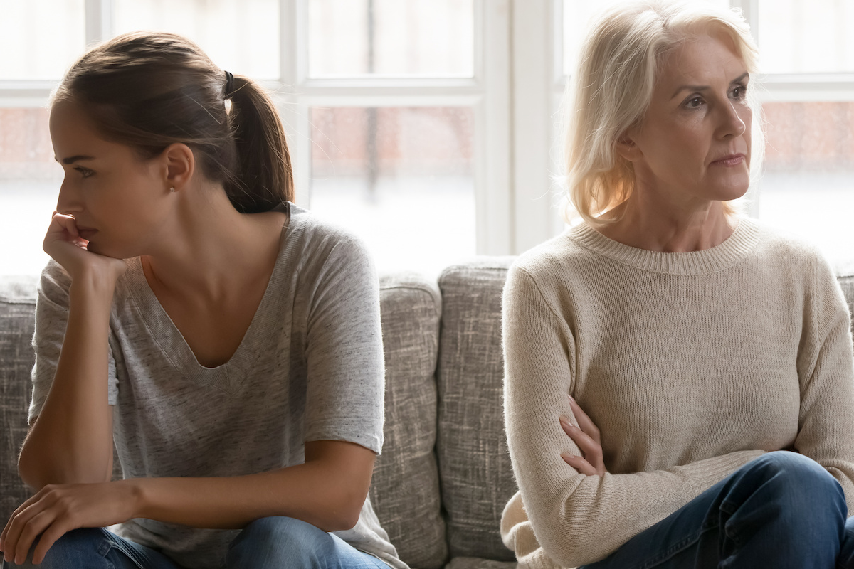 Adult daughter and mother sitting apart on a couch, both looking away from each other with tense expressions