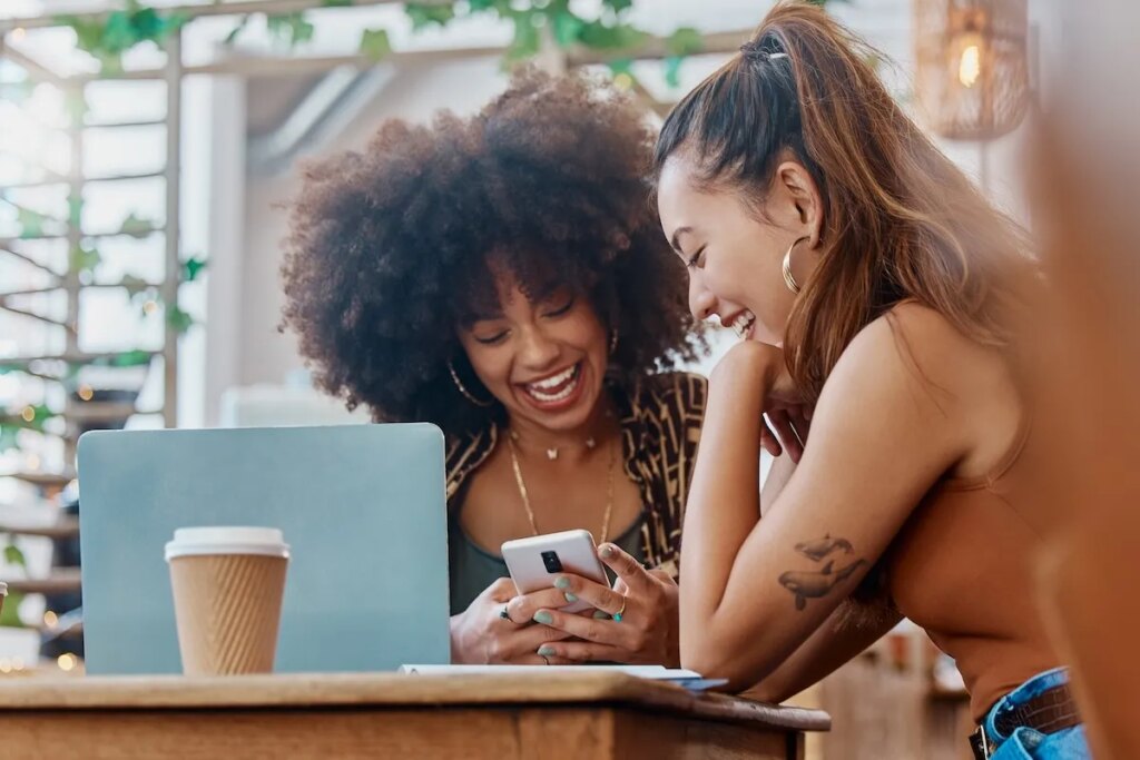 Two women sitting at a coffee shop, laughing and looking at a phone
